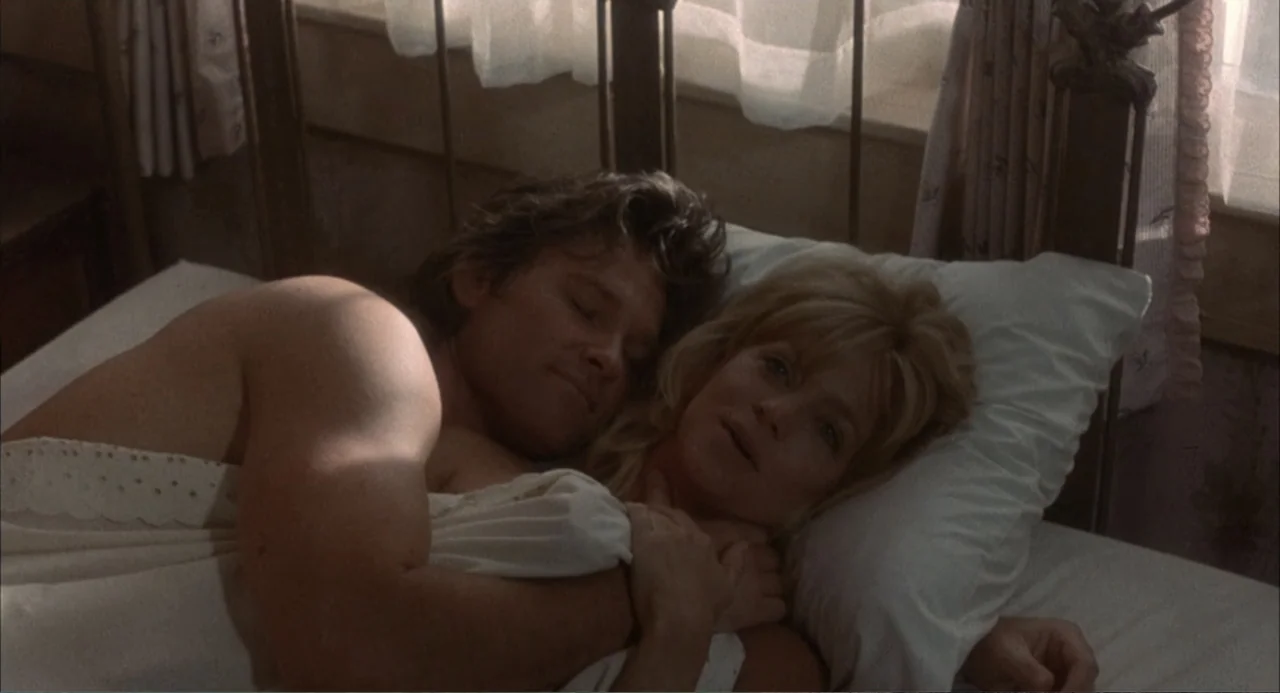 Overboard (1987) movie still: medium — A sunlit bedroom features a metal bed frame covered by white bedsheets. A man and a woman lie together…