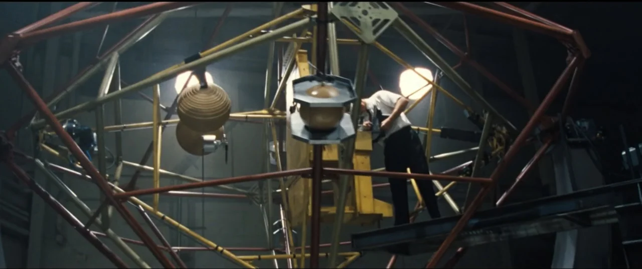 First Man (2018) movie still: medium — A dimly lit industrial space features a massive complex red and yellow metal framework. A man in a white…