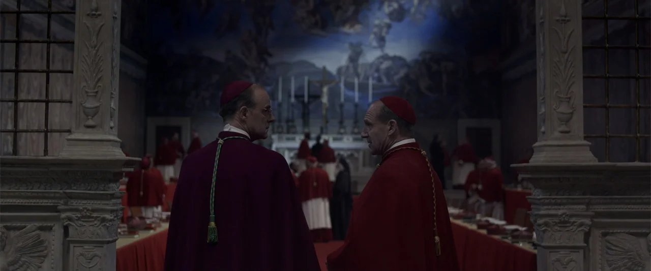 Conclave (2024) movie still: medium — Two men in red ecclesiastical vestments stand in a dimly lit ornate hall. They face each other while other…