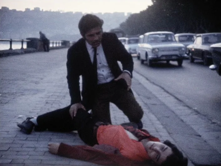 Eika Katappa (1969) movie still: fullbody — A stone-paved walkway runs parallel to the sea during a grey evening. A man in a dark suit kneels over a…