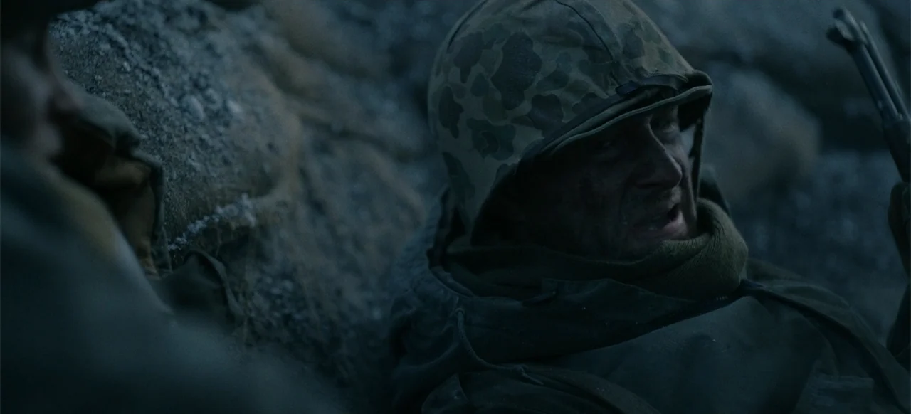 Devotion (2022) movie still: closeup — A muddy, dark foxhole during a daytime combat operation. A soldier in a camouflage helmet pants with exertion…