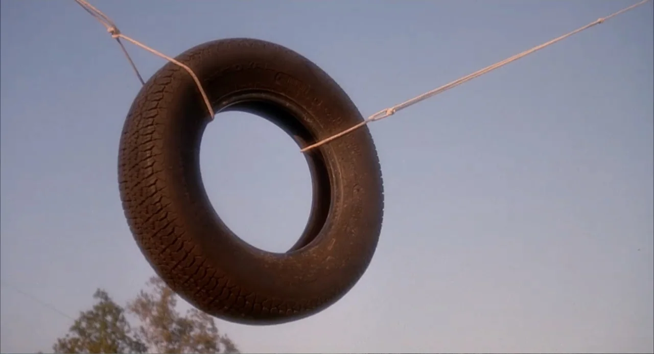 Teen Witch (1989) movie still: closeup — A worn black rubber tire hangs from two ropes against a clear blue sky. The tire is suspended outdoors with…
