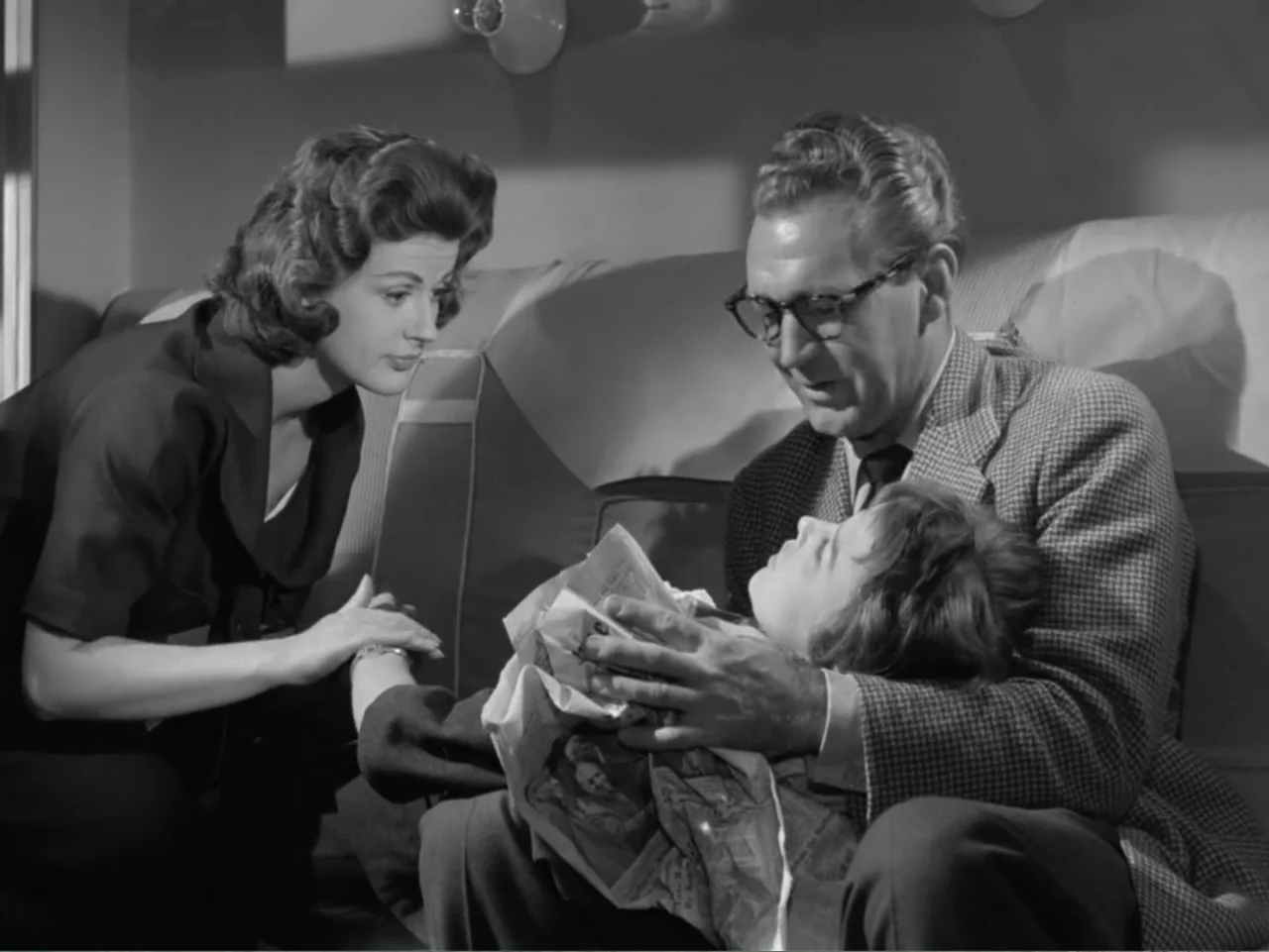 The Crawling Eye (1958) movie still: medium — A dimly lit airplane cabin with upholstered seats and overhead reading lights. A man comforts a…