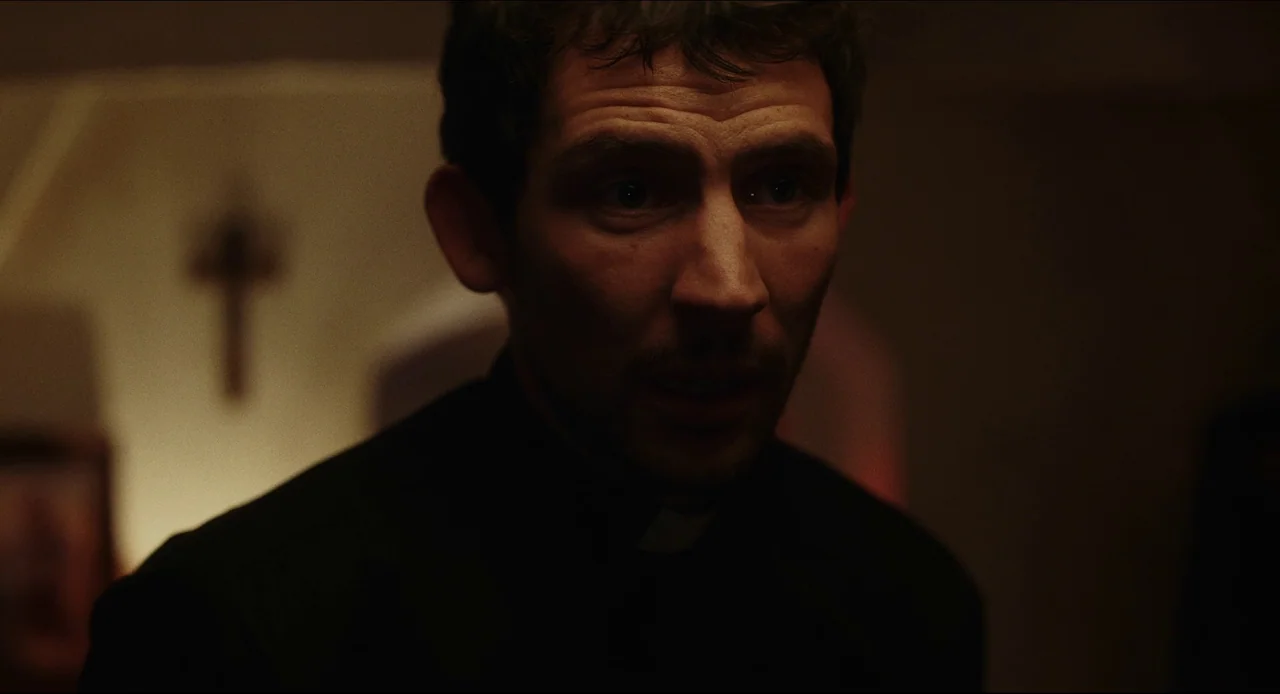 Wake Up Dead Man (2025) movie still: closeup — A dimly lit room features a small wooden crucifix mounted on a beige wall. A man in clerical attire…