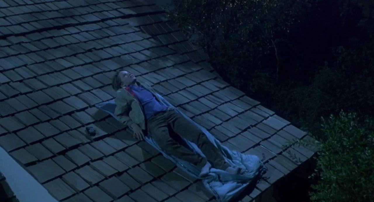 Explorers (1985) movie still: wide — A steeply pitched wooden shingled roof is set against a dark night background. A teenager lies on a blue…