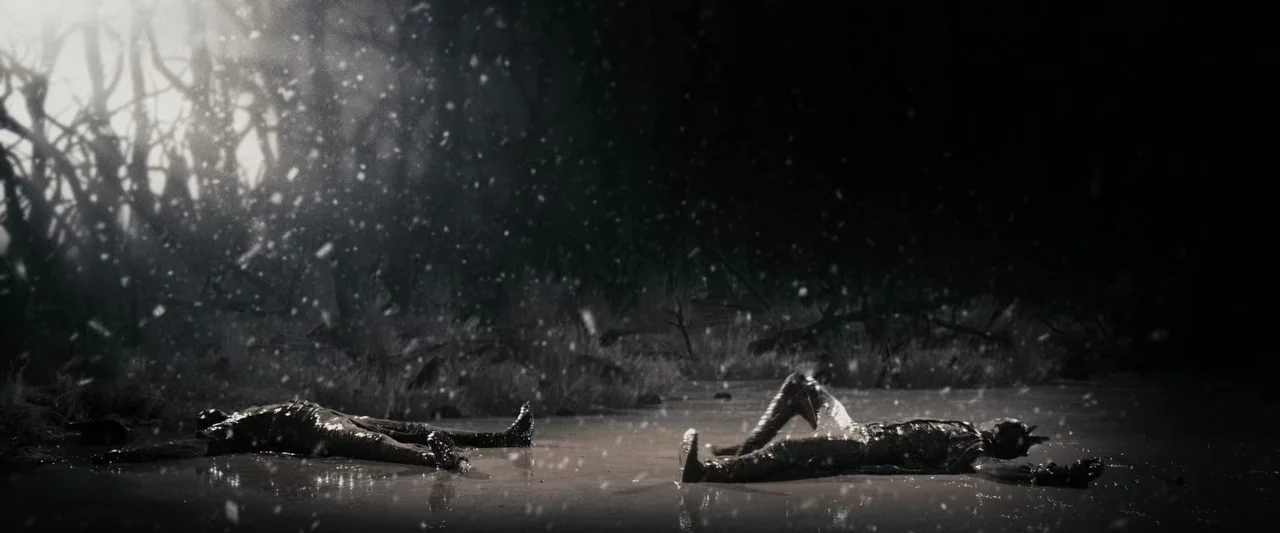 The Spirit (2008) movie still: wide — A dark muddy area at night features falling snow backlit by a bright light. Two figures lie motionless on the…