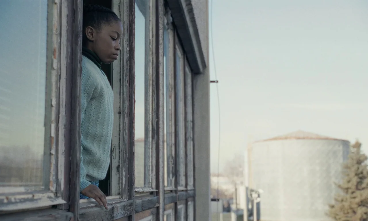 The Silent Twins (2022) movie still: medium — A young girl leans out of an old wooden window frame in a building. She looks out towards a distant…