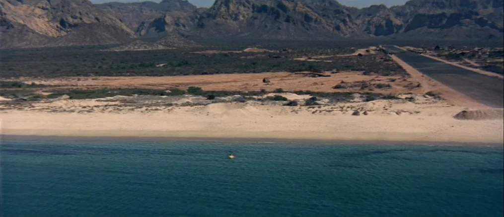Catch 22 (1970) movie still: establishing — A vast coastal landscape features a sandy beach and a road meeting dry mountains. A lone yellow object…