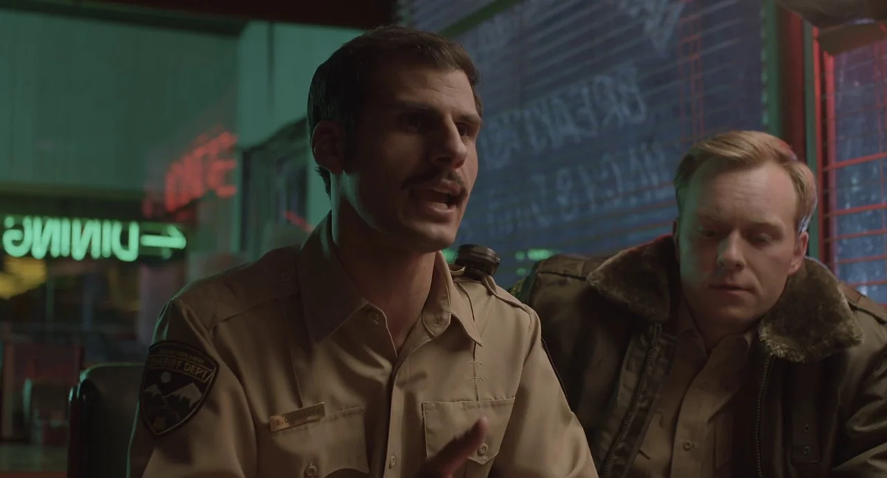 The Wolf of Snow Hollow (2020) movie still: medium — Two men sit together in a booth inside a dimly lit diner at night. One man in a uniform speaks…