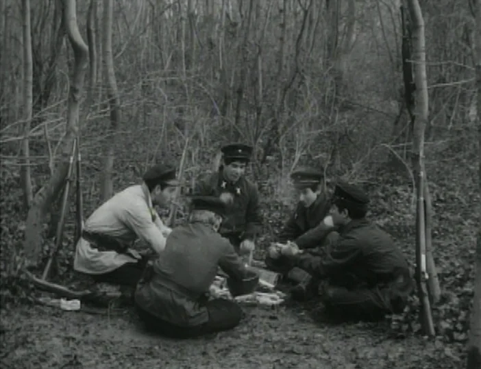 Les Carabiniers (1963) movie still: medium — A group of soldiers sits in a circle amidst bare trees in a woodland. They are gathered around a…