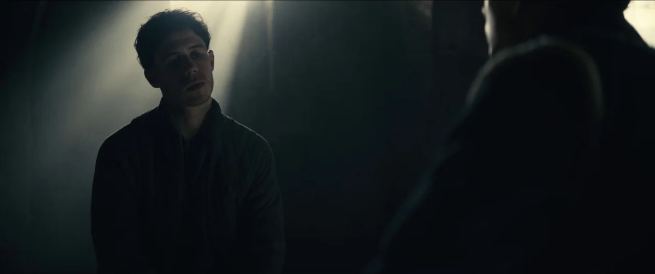 The King movie still: medium — A dimly lit room features a single beam of light piercing through darkness. A young man in a dark jacket sits facing…