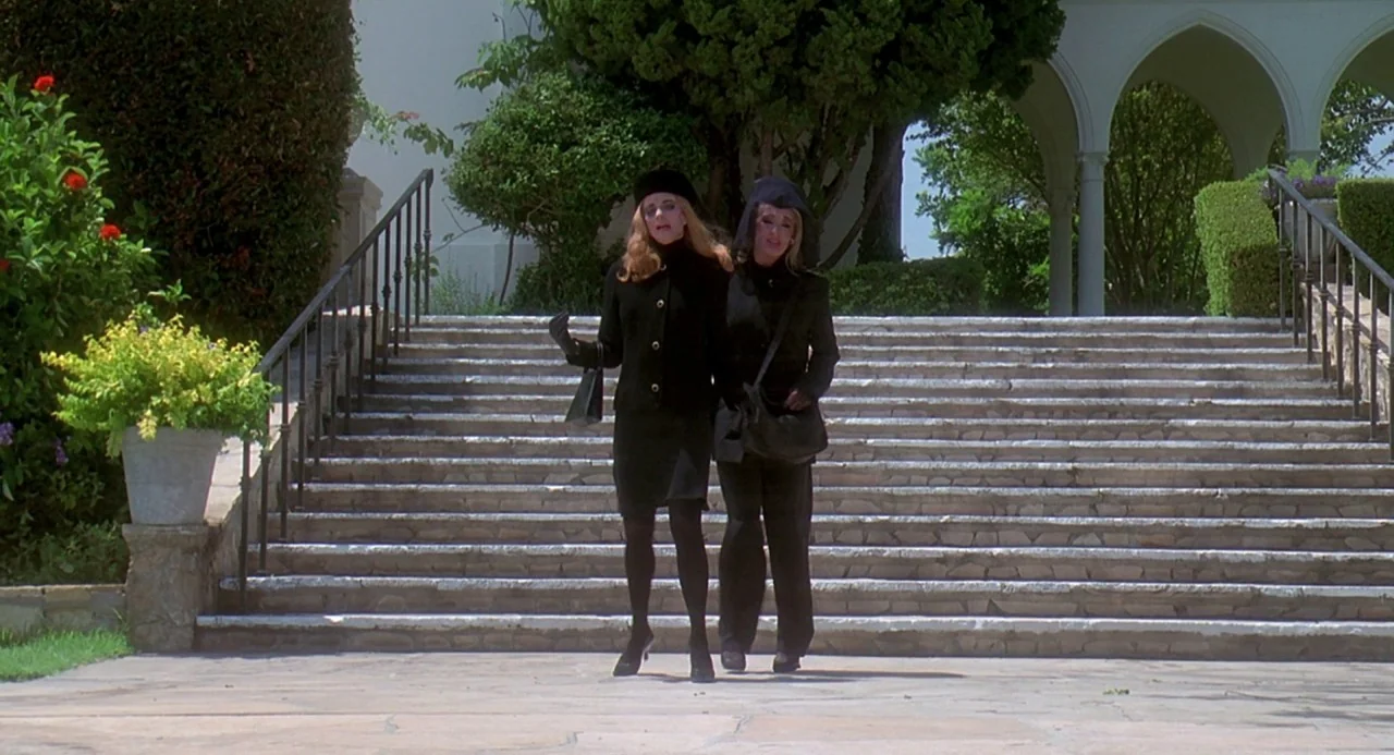 Death Becomes Her (1992) movie still: fullbody — Two women dressed in black mourning attire stand together on wide stone steps. Lush greenery and…