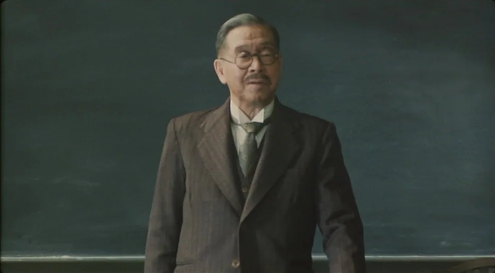 Madadayo (1993) movie still: medium — A dark, blank blackboard serves as the background in an old-fashioned classroom setting. An elderly man in a…