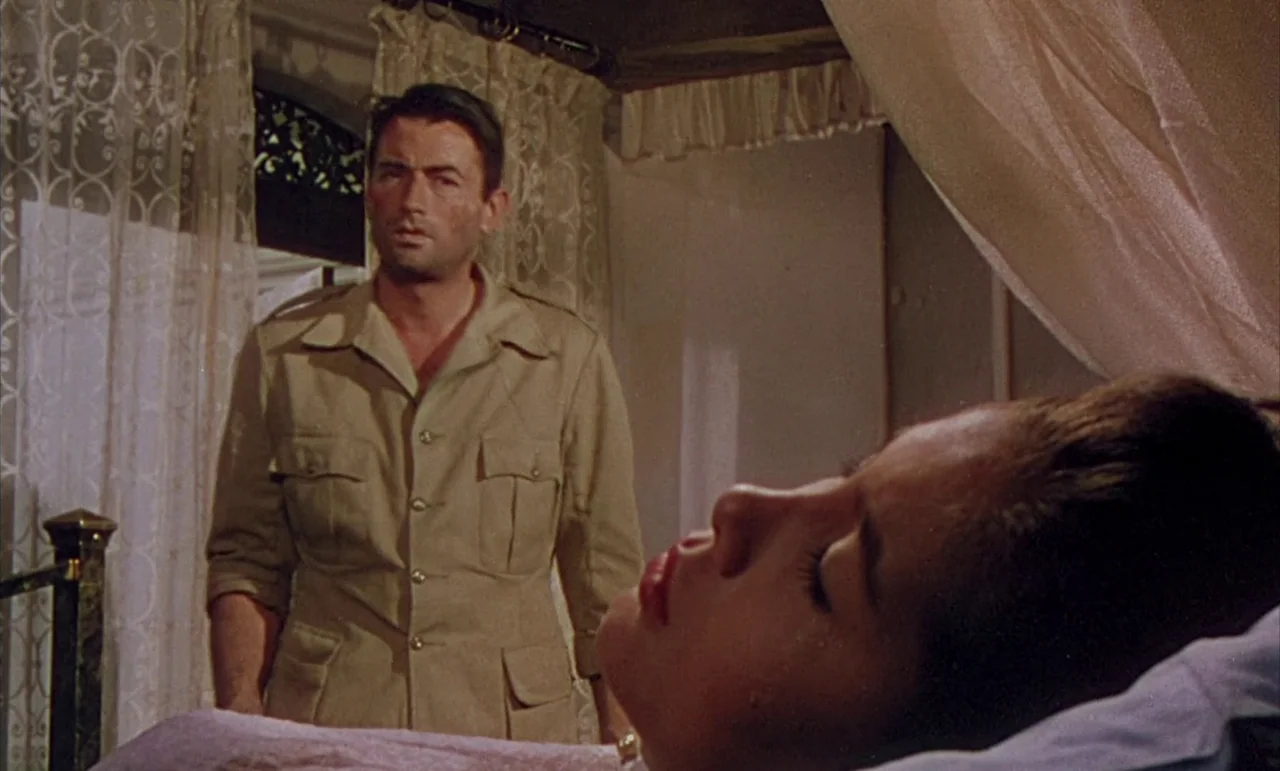 The Purple Plain (1954) movie still: medium — An interior bedroom features a man standing before a woman lying on a bed. Lace curtains cover the…