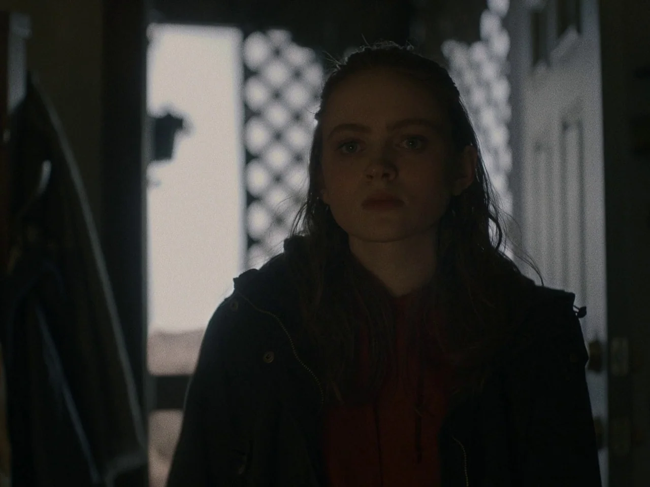 The Whale (2022) movie still: medium — A young girl stands in a dark indoor threshold facing the camera. Bright light spills into the room from the…