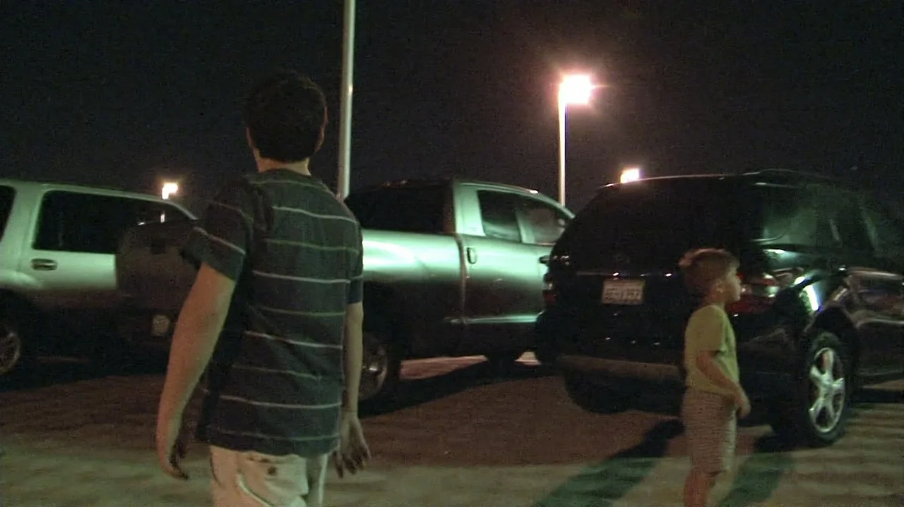 Homeless: The Motel Kids of Orange County (2010) movie still: fullbody — A nighttime parking lot features several dark parked vehicles under bright…