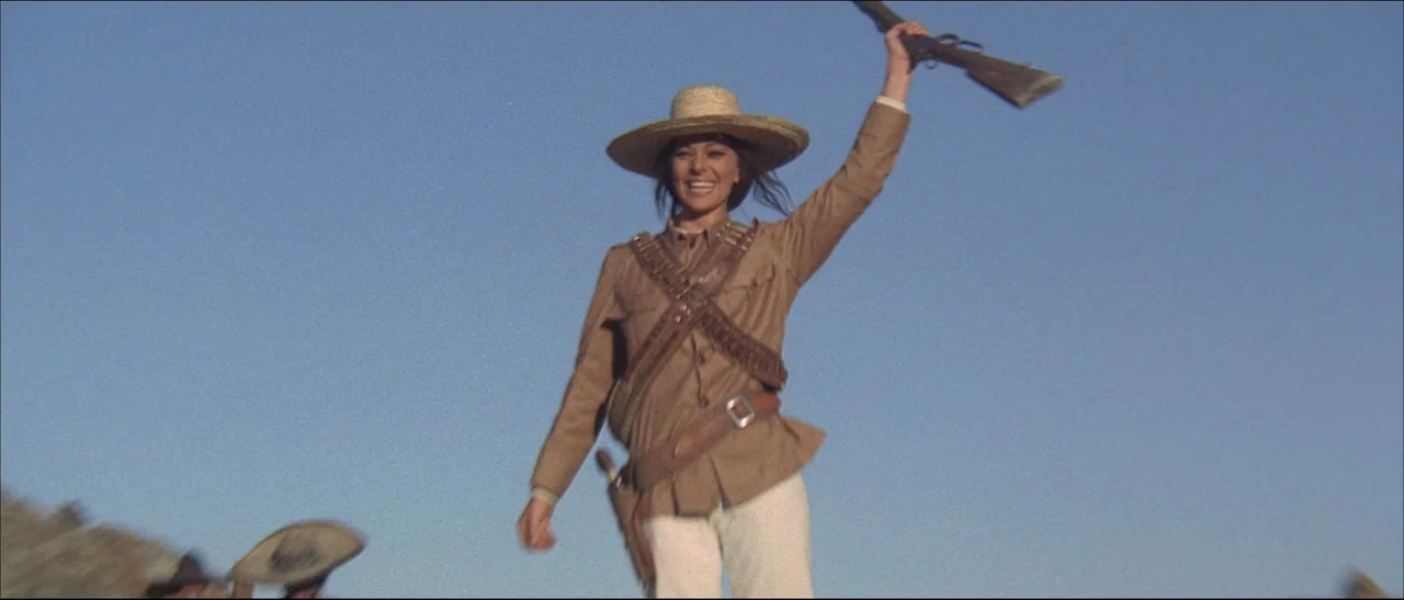 The Mercenary (1968) movie still: fullbody — A woman stands under a clear blue sky in an arid, rocky desert landscape. She smiles broadly while…