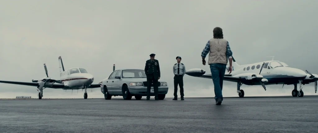 MacGruber (2010) movie still: wide — An overcast airfield features two small private planes parked flanking a sedan and three men. A man approaches…