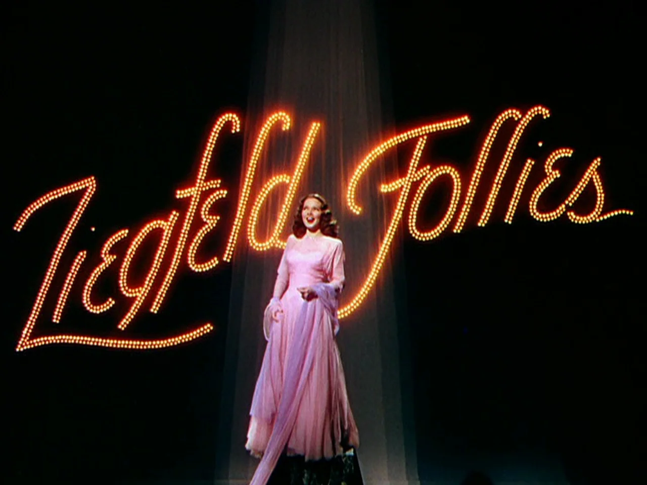 Ziegfeld Follies (1945) movie still: fullbody — A dark theatrical stage features a massive neon sign reading Ziegfeld Follies. A woman stands…