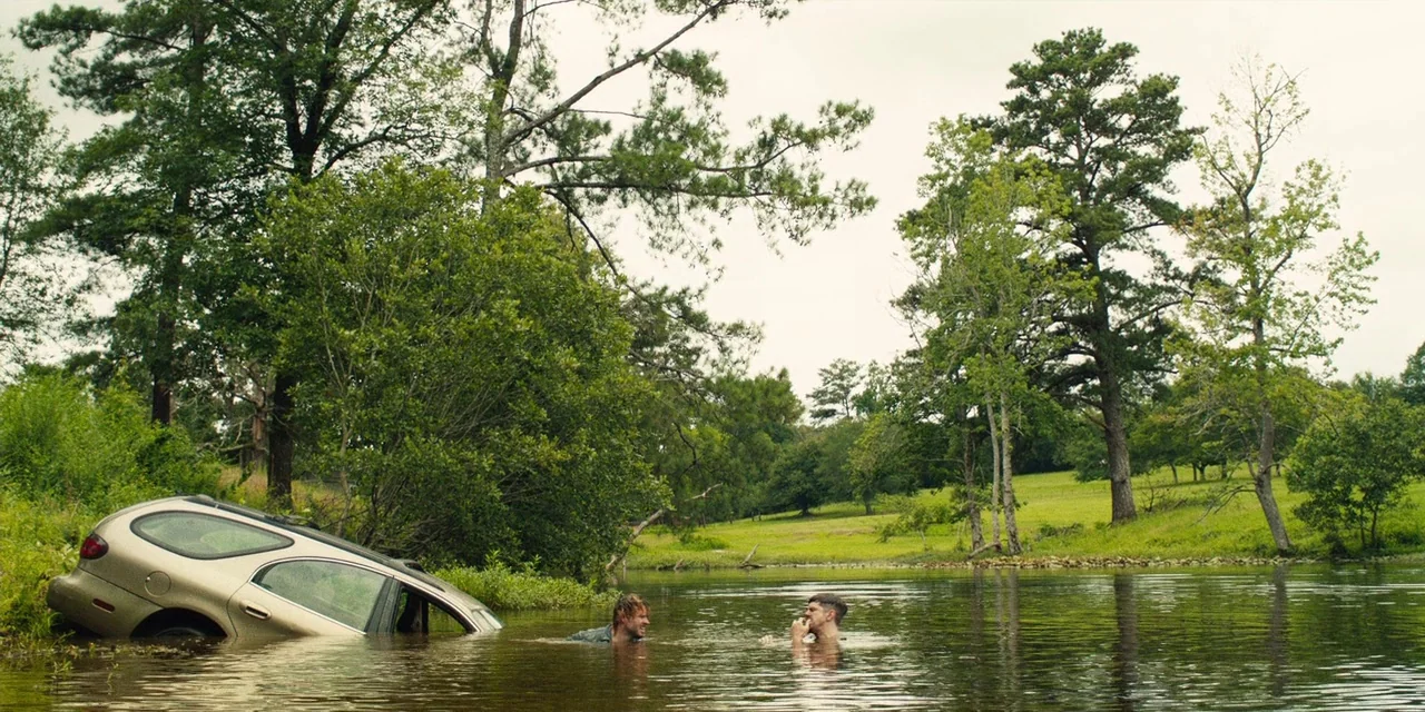 The Death of Dick Long (2019) movie still: wide — A rural pond with a partially submerged silver station wagon near the bank. Two men stand in the…