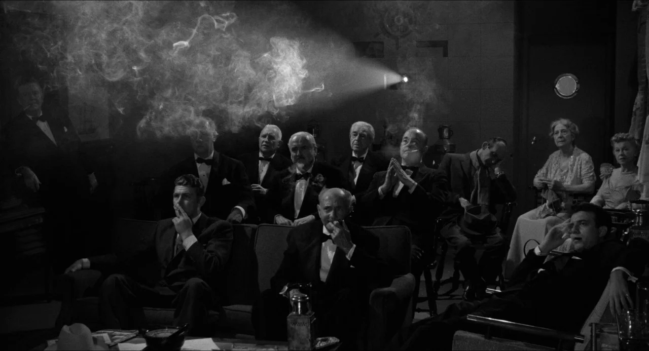 A Face In The Crowd (1957) movie still: wide — A dimly lit room functions as a private screening area filled with dense tobacco smoke. A group of…
