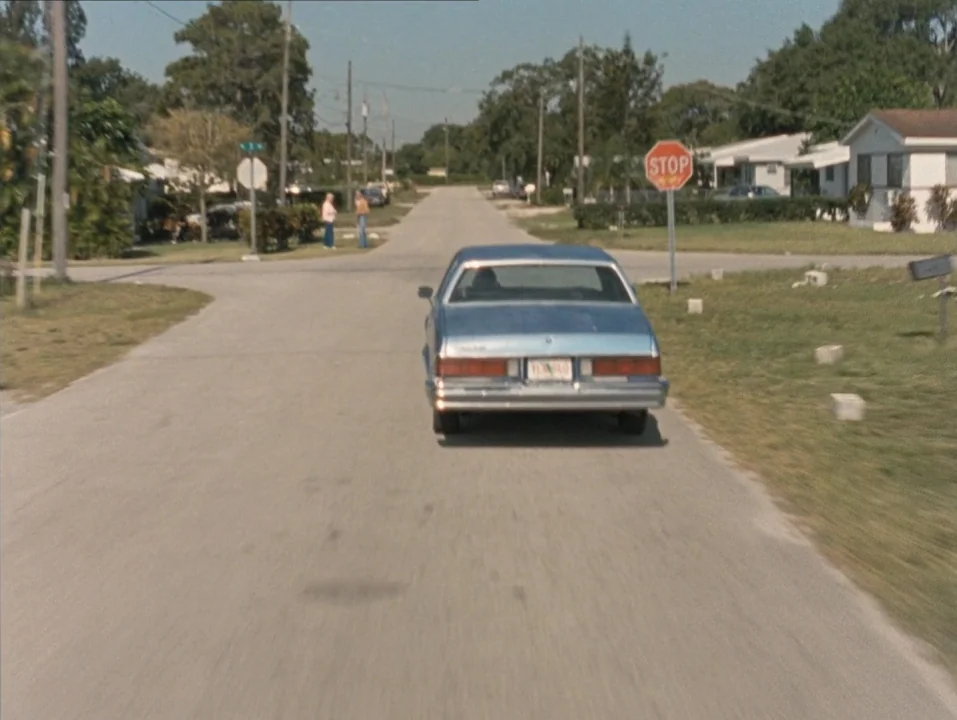 River of Grass (1994) movie still: wide — A quiet residential intersection features a lone sedan moving away down a paved road. Two distant figures…