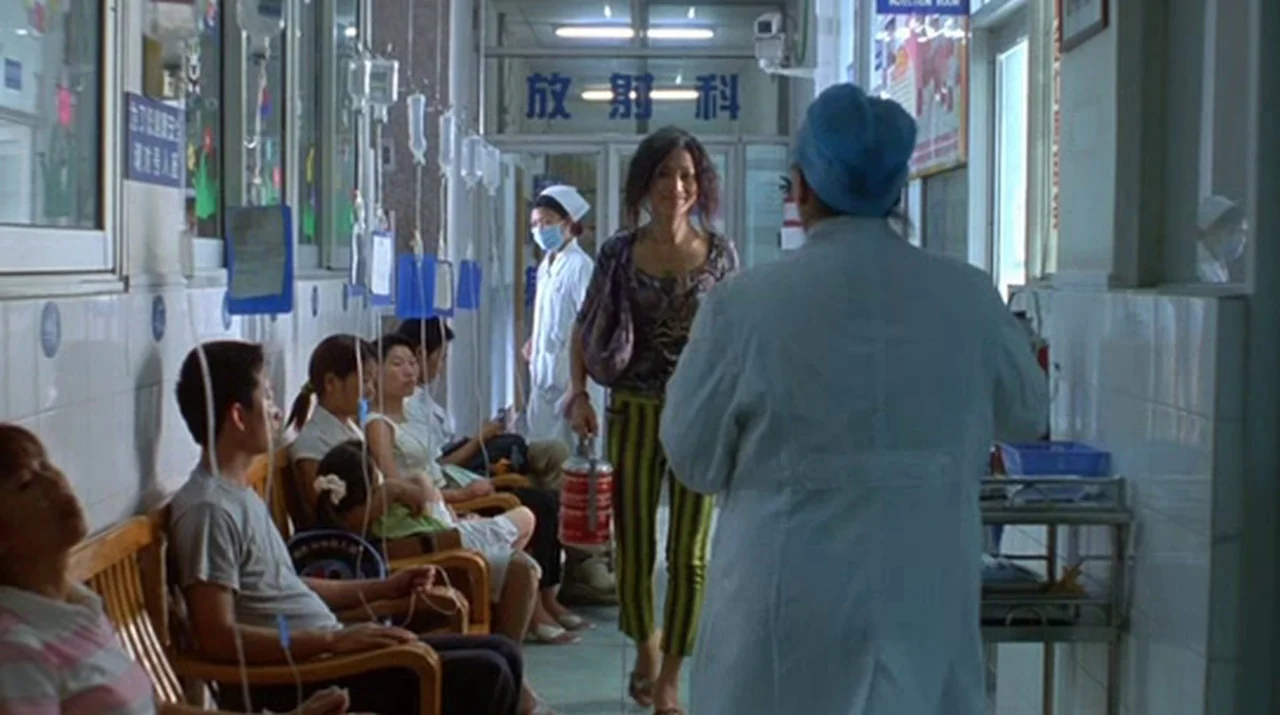 Dumplings (2004) movie still: medium — A hospital hallway features tiled walls and multiple patients sitting in wooden chairs receiving IVs. A woman…