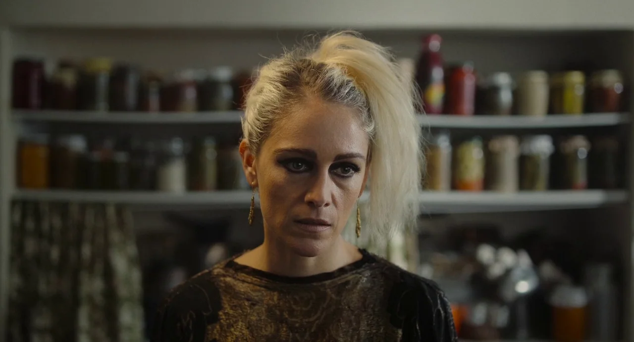 Flux Gourmet (2022) movie still: medium — A pantry shelf filled with various glass jars containing preserved foods is visible. A woman with bleach…