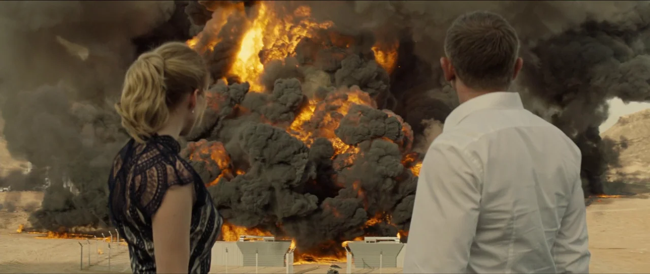 Spectre (2015) movie still: wide — A vast desert location with a large fiery explosion rising into the air. A woman and a man stand in the…