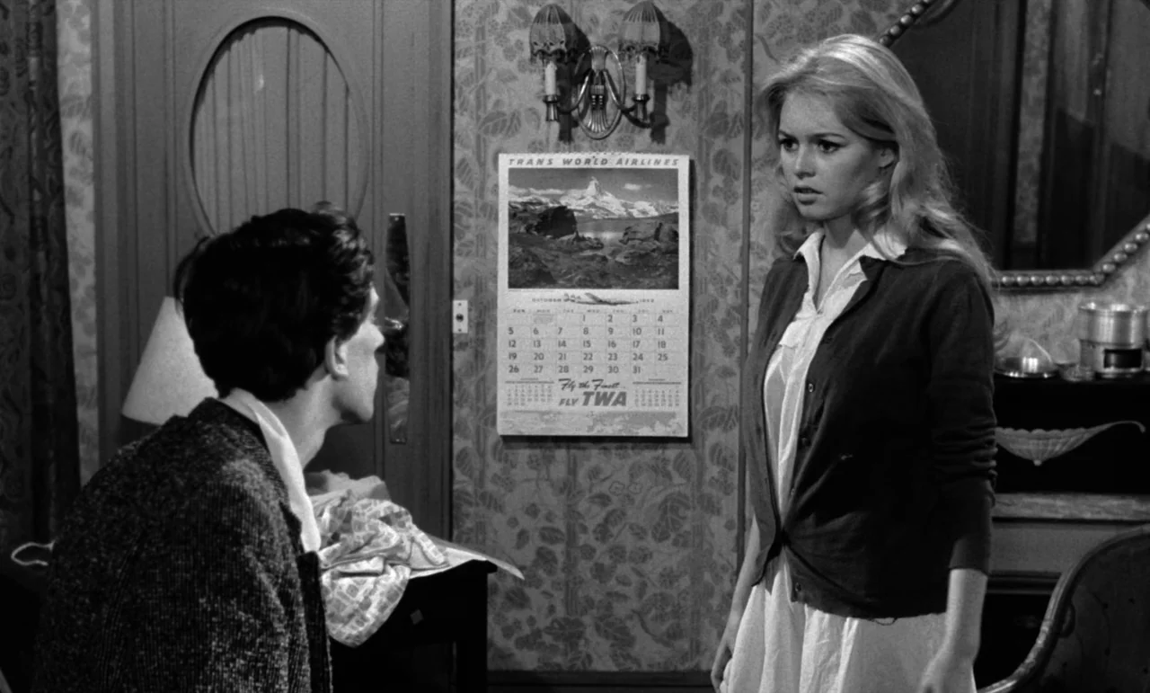 La Verite (1960) movie still: medium — A cluttered room features patterned wallpaper and a wall-mounted TWA travel calendar. A woman in a light…