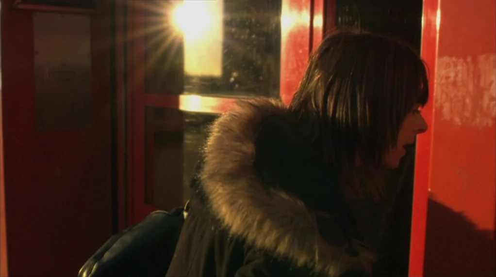 Red Road (2006) movie still: medium — A person stands inside a bright red phone booth at night under dark conditions. The subject looks through the…