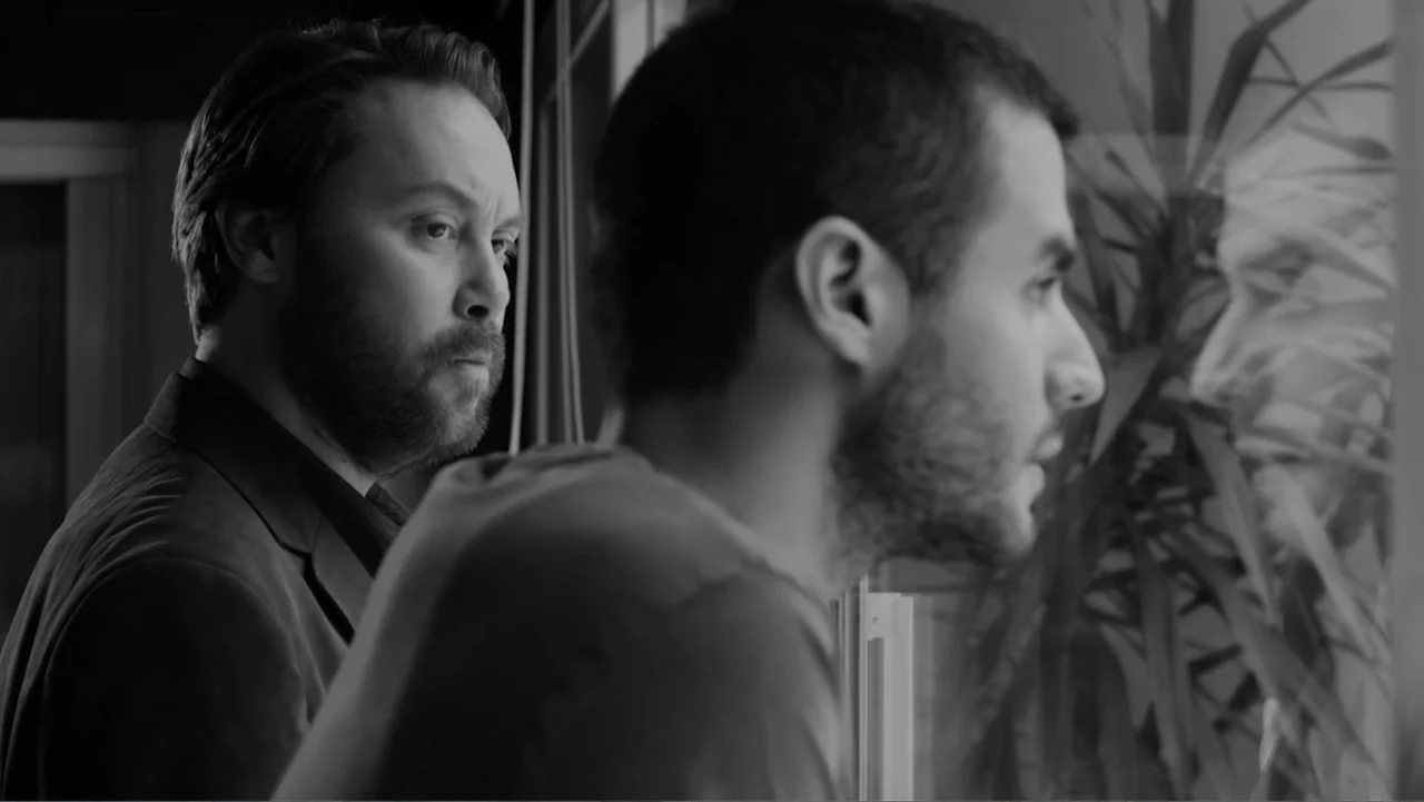Creditors (2025) movie still: medium — A dark indoor space with a window looking out onto a plant. Two men stand together, one closer to the glass…