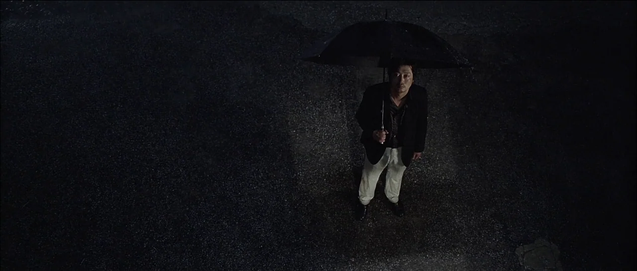 The Chaser (2008) movie still: fullbody — A dark, wet asphalt surface reflects dim light during a rainy night. A man stands alone holding a black…
