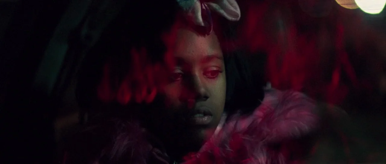 Good Time (2017) movie still: closeup — A young girl sits inside a dark vehicle at night illuminated by red light. Reflections of red light obscure…