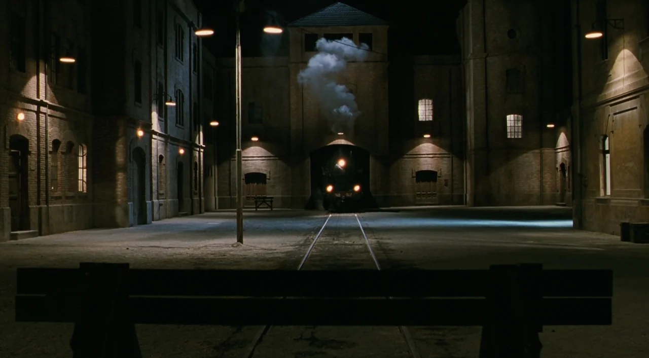 Life is Beautiful (1997) movie still: wide — A stone train depot courtyard at night is illuminated by rows of streetlamps. A dark steam locomotive…