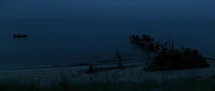 Jaws movie still: wide — A dark, calm beach scene at blue hour features a wooden pier extending into water. A small boat drifts on the horizon near…