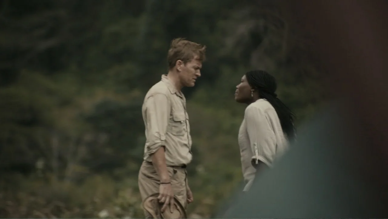 The Dinosaur Project (2012) movie still: medium — A dense green natural environment serves as the backdrop for two people conversing. A man and a…