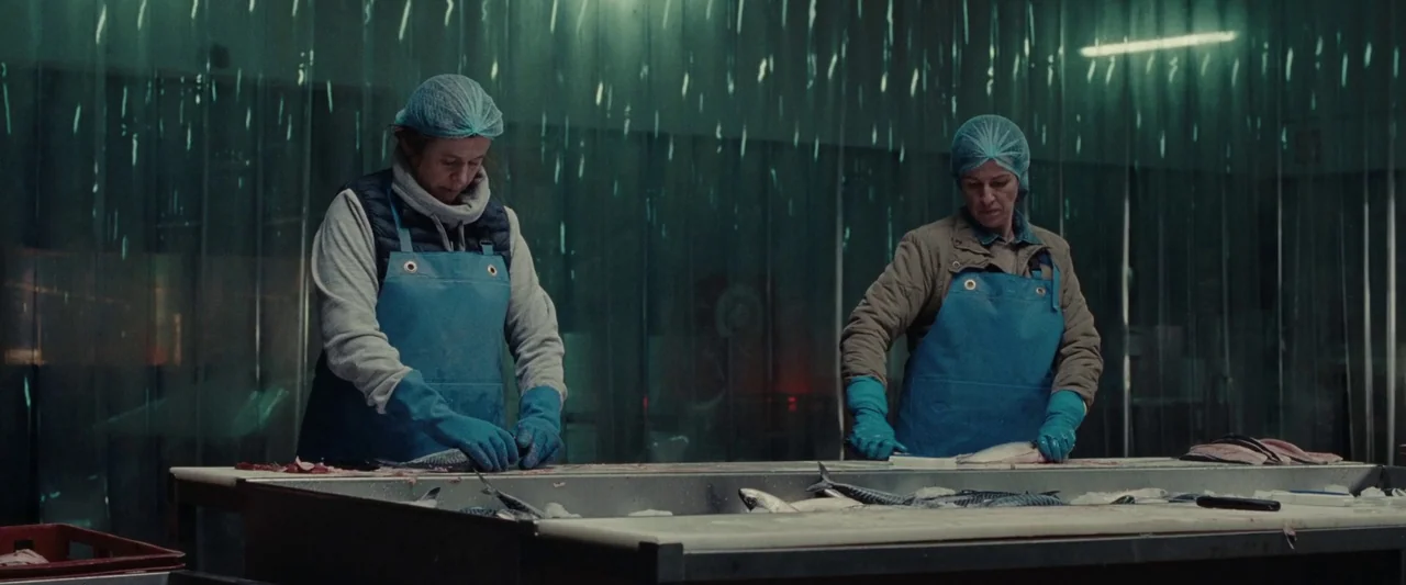 God’s Creatures (2022) movie still: medium — Two women stand at a stainless steel table in a sterile fish processing room. They are working…