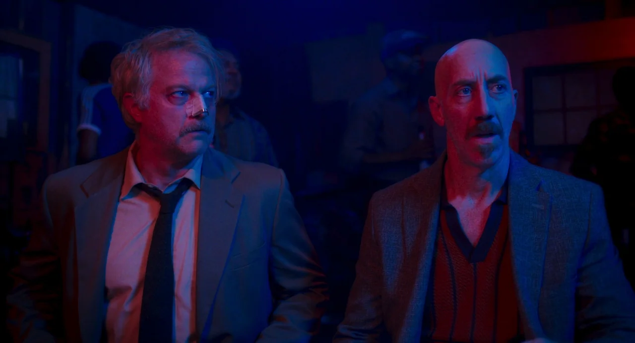 Drive-Away Dykes (2024) movie still: medium — A dark, crowded venue with patrons silhouetted in the background under neon lighting. Two men sit in…