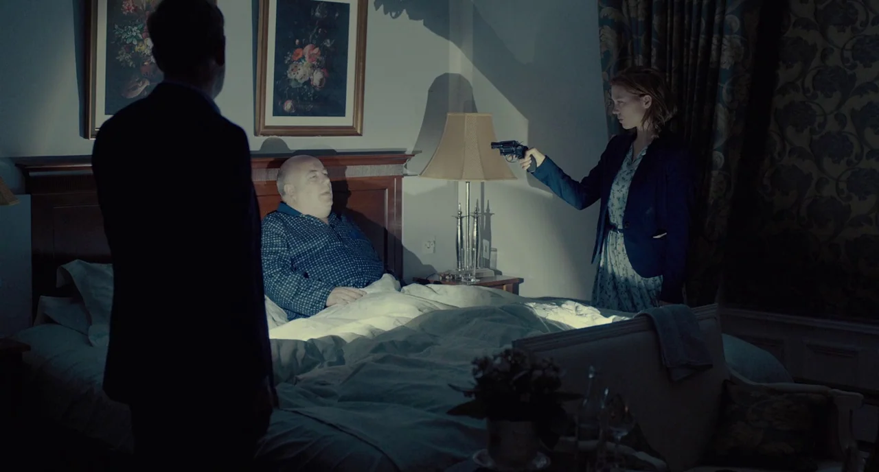 The Lobster (2015) movie still: wide — A dimly lit hotel room features floral wallpaper and framed floral artwork above a bed. A woman aims a…