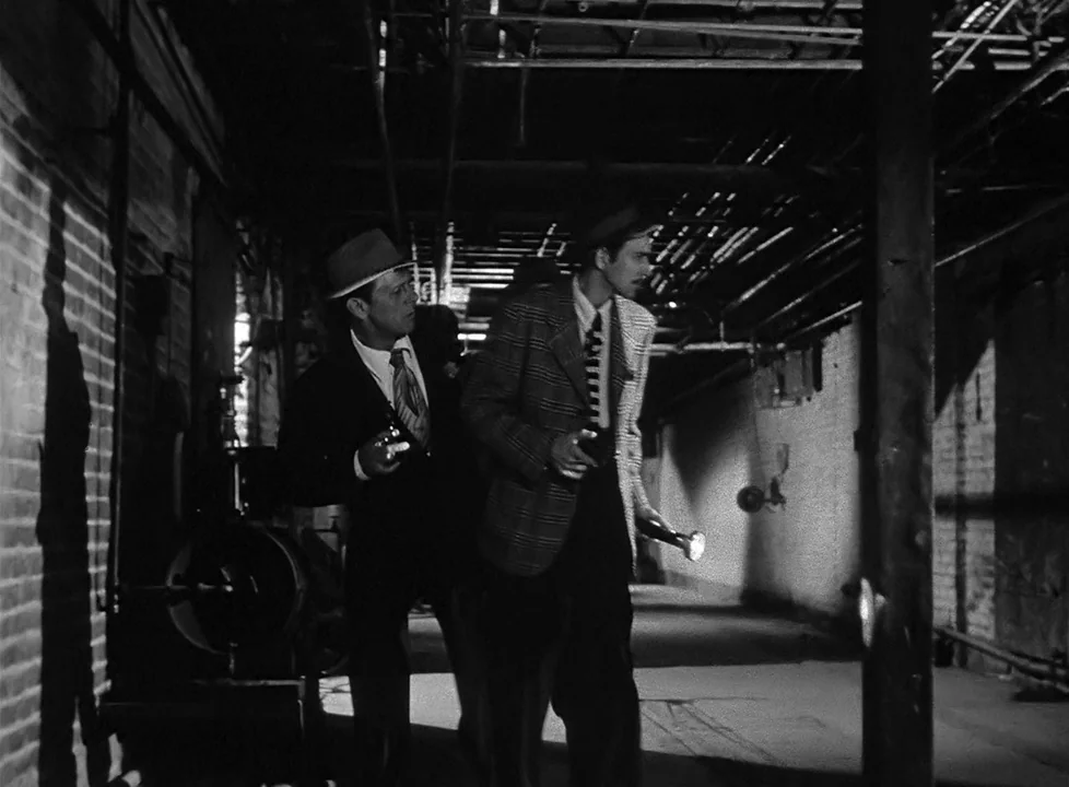M (1951) (1951) movie still: fullbody — A long industrial corridor features exposed ceiling pipes and brick walls under dim light. Two men in suits…