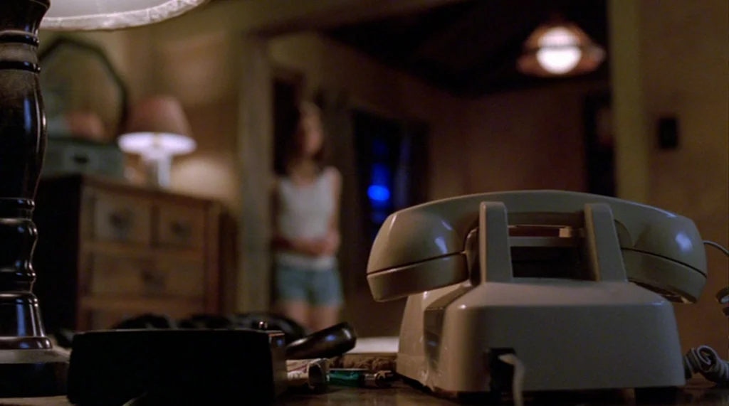 Bug movie still: closeup — A cream-colored vintage landline telephone rests on a desk in a dark room. The background shows a dimly lit blurred…