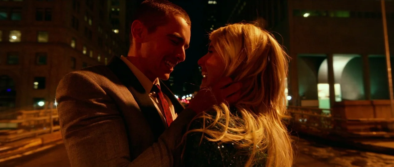 Nerve (2016) movie still: medium — A man and a woman embrace on a bridge at night with city lights behind. They stand close together smiling while…