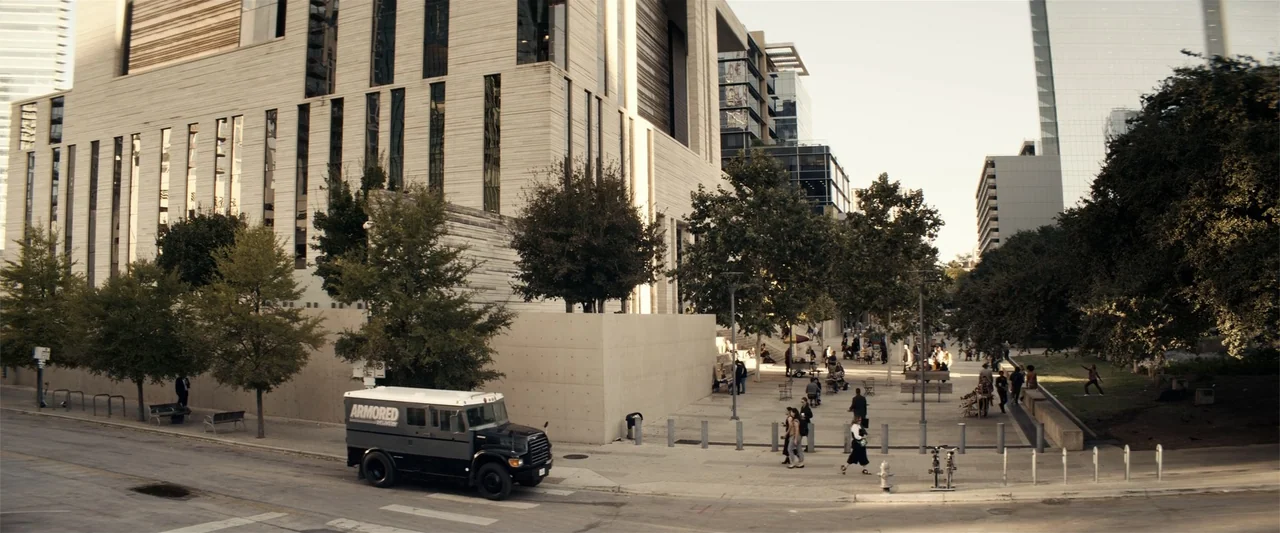 Hypnotic (2023) movie still: wide — A modern urban plaza features a large stone building and surrounding trees and walkways. A black and grey…