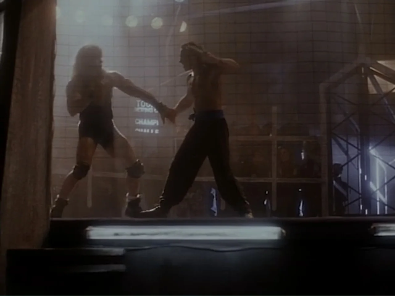 Firepower (1979) movie still: fullbody — A dimly lit industrial style fighting ring with metal cage fencing in the background. Two shirtless men in…