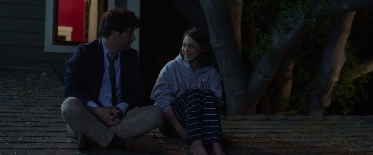 Night Owls (2015) movie still: medium — A dark, shingled rooftop at night with a large tree trunk visible nearby. A man and a girl sit side-by-side…
