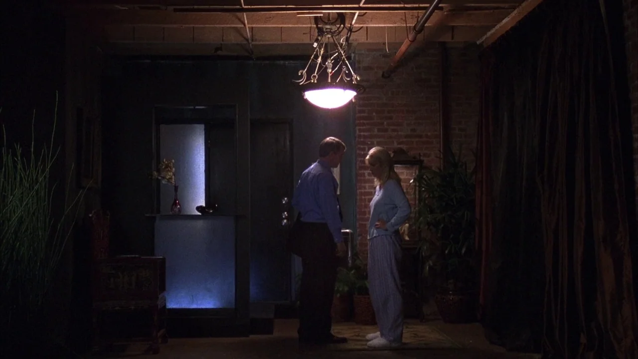 Saw movie still: wide — A basement with dark brick walls, exposed ceiling joists, and heavy curtains to one side. A man and a woman stand close…