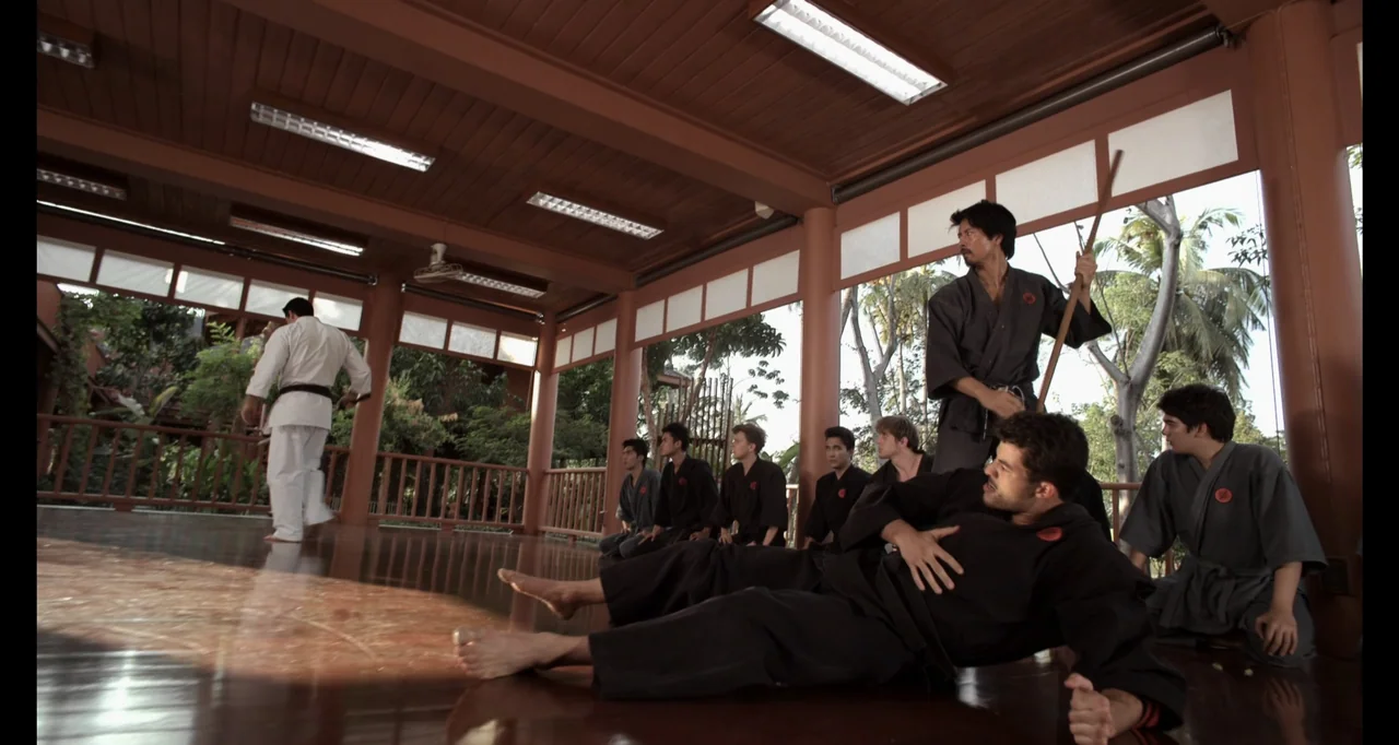 Ninja: Shadow of a Tear (2013) movie still: wide — A spacious wooden dojo with open sides looks out onto tropical palm trees. A group of students in…