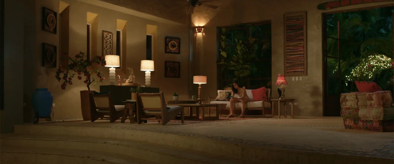 Beautiful Wedding (2024) movie still: wide — A spacious living room features rattan furniture and soft warm lighting at night. A woman sits on a…