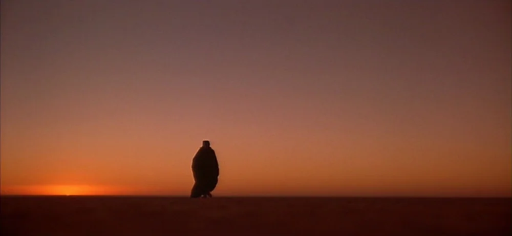 Lawrence Of Arabia movie still: wide — A vast, barren desert landscape stretches to the flat horizon under a sunset sky. A solitary hooded figure…